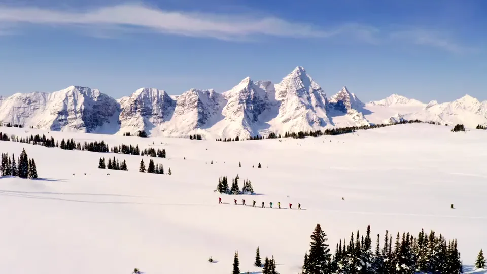 A line of ski tourers crossing a wide snowfield below a wall of snow-covered peaks, seen from the air