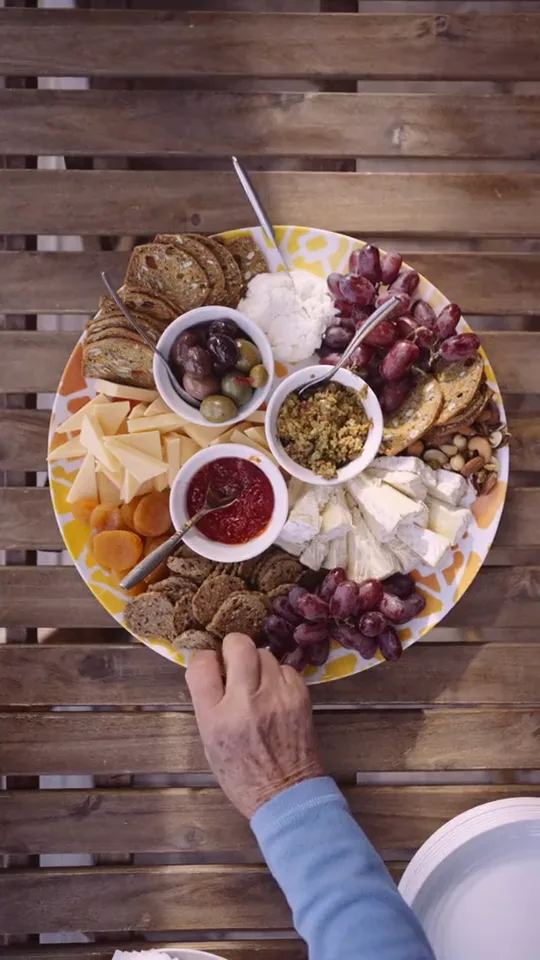 A round board of cheeses, crackers, grapes, dried fruit, nuts and dips set down on the timber table from above