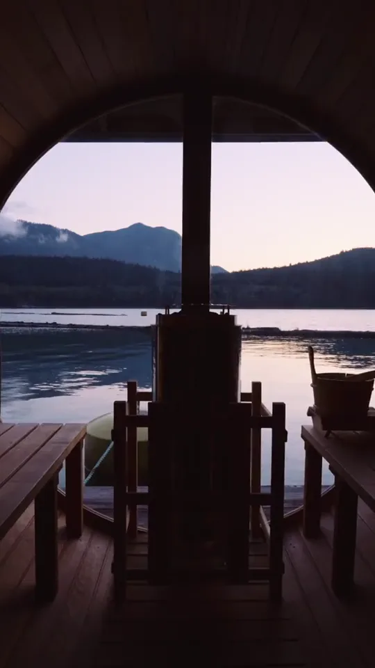 Inside the barrel sauna at dusk, the stove and bench in silhouette against the round window and the pink inlet beyond