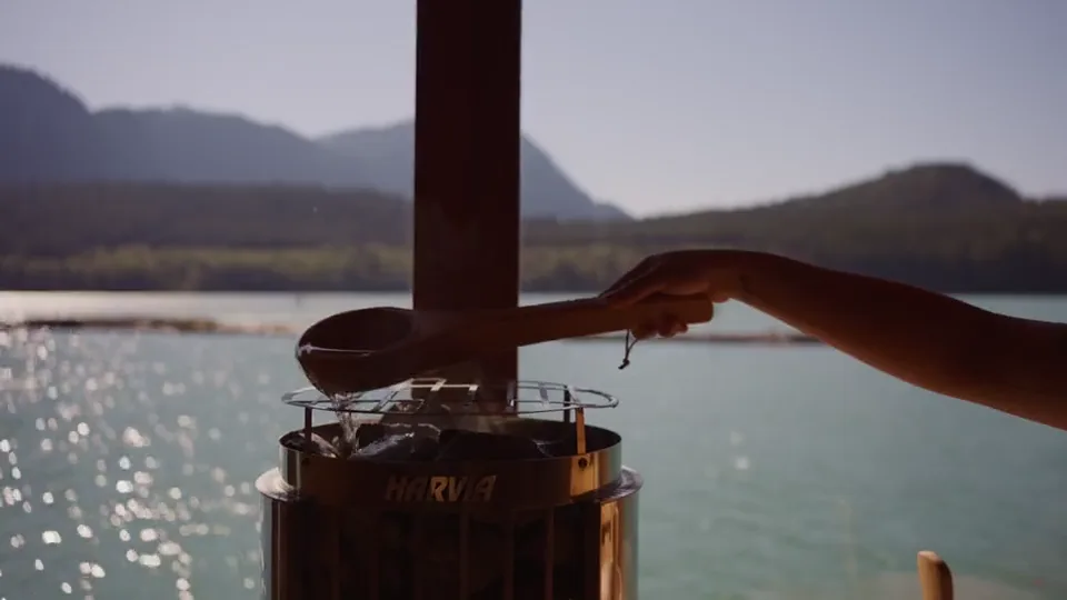 A hand ladling water onto the sauna stove's rocks, the inlet bright through the window behind