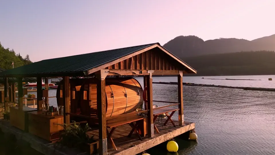 A cedar barrel sauna under a small roof on Knight Inlet Lodge's floating dock at sunset, the inlet and mountains behind it turning pink
