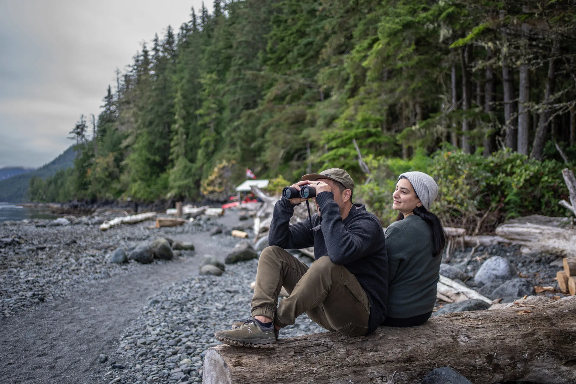 Two guests sitting back to back on a driftwood log on a pebble beach, one in a ball cap looking through binoculars, the other in a grey wool beanie