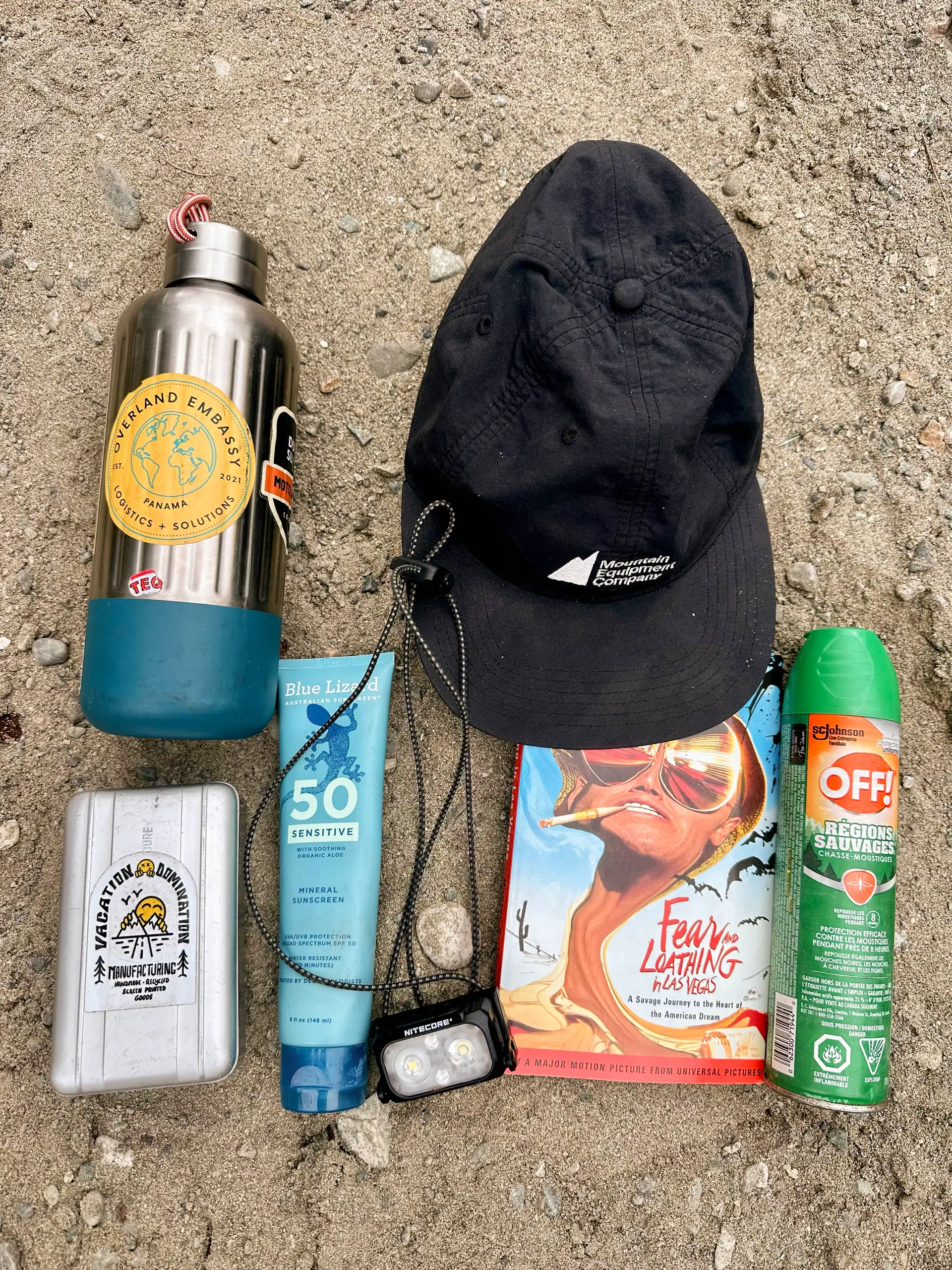 Packing items laid out on grey gravel: a steel water bottle, a black cap, a tube of mineral sunscreen, a headlamp, a battery pack, a can of insect repellent and a paperback
