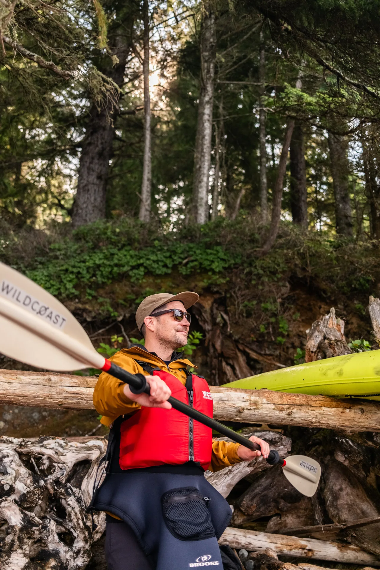 A paddler in a mustard rain jacket, red life jacket and cap holding a Wildcoast paddle across his chest in front of driftwood and a green kayak
