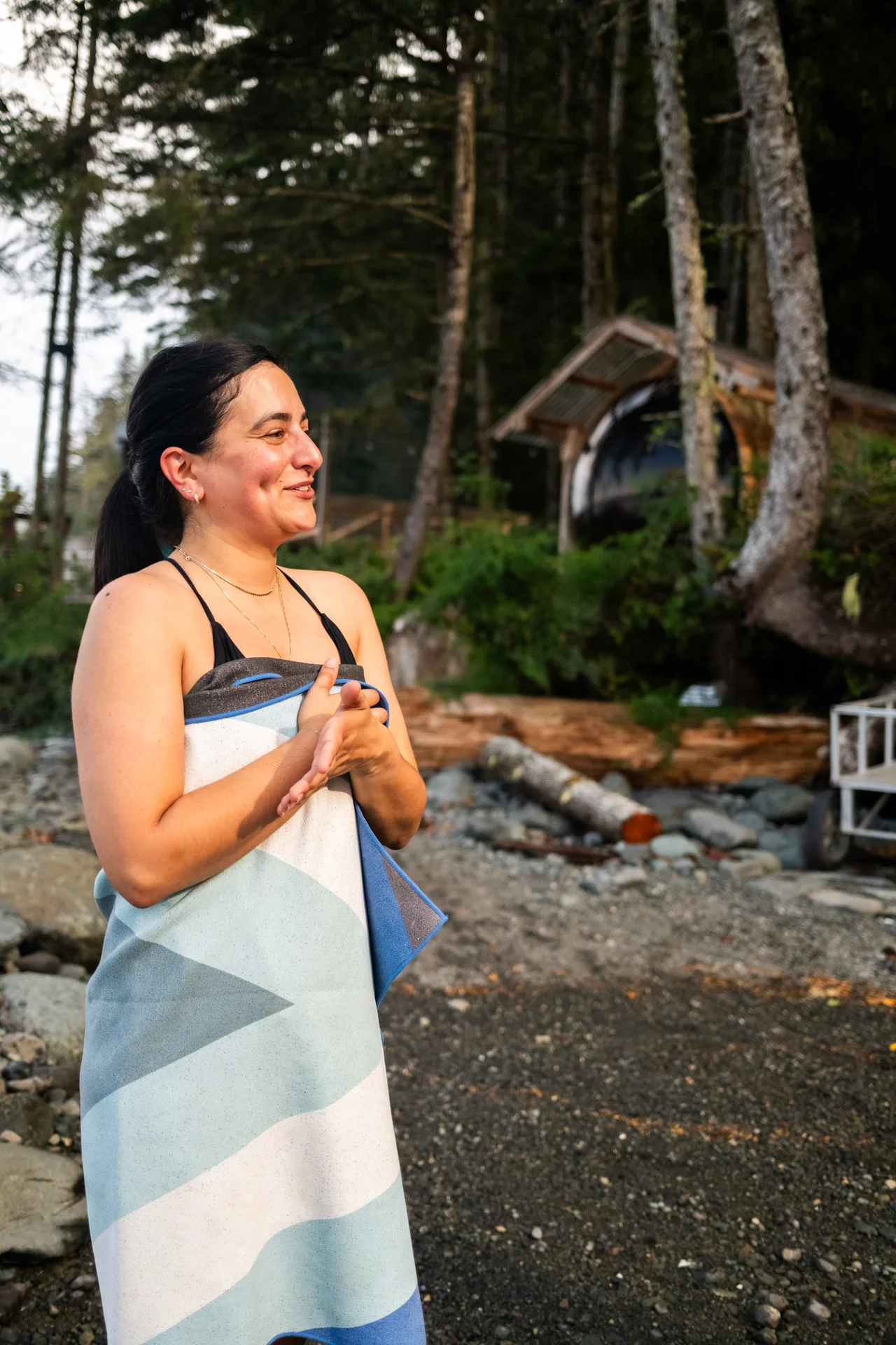 A guest on a pebble beach wrapped in a fast-dry towel after a swim, with the camp sauna among the trees behind her