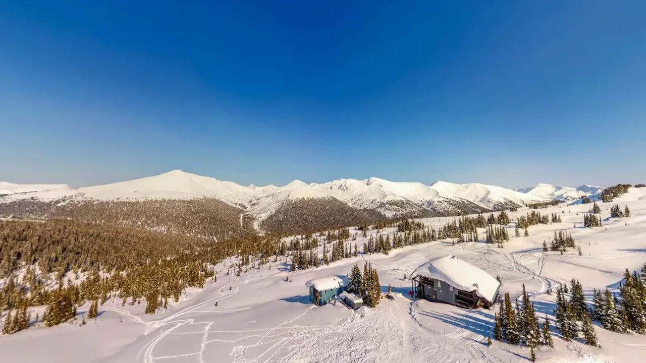 Purcell Mountain Lodge and its chalet on the winter bench from above, ski tracks radiating across the snow toward the treeline and the peaks beyond